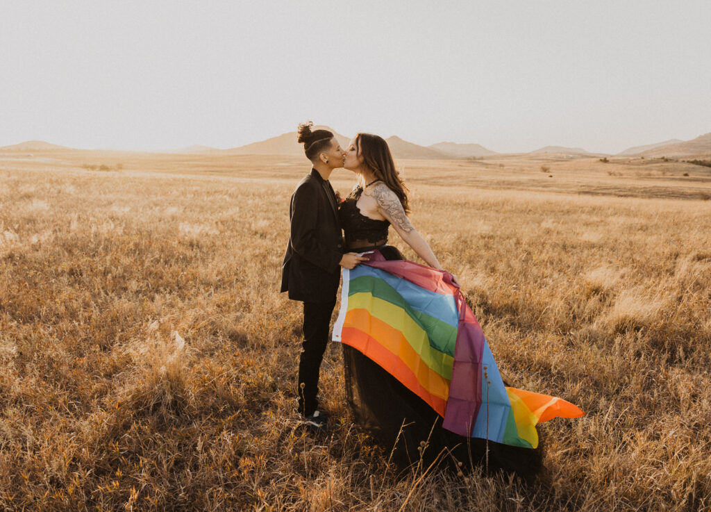 A queer couple stands together kissing and holding a rainbow flag on their elopement day in the Wichita mountains of Oklahoma.