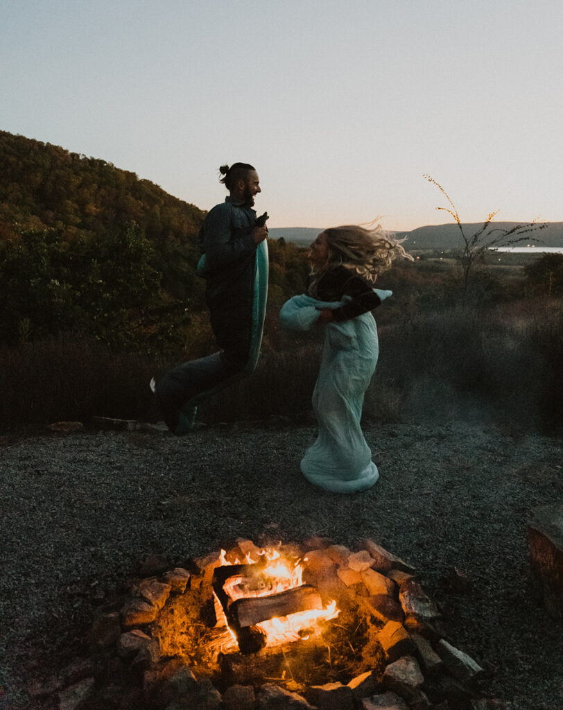 A couple jumping in their sleeping bags at night by a fire to celebrate their elopement day in Broken bow, oklahoma.