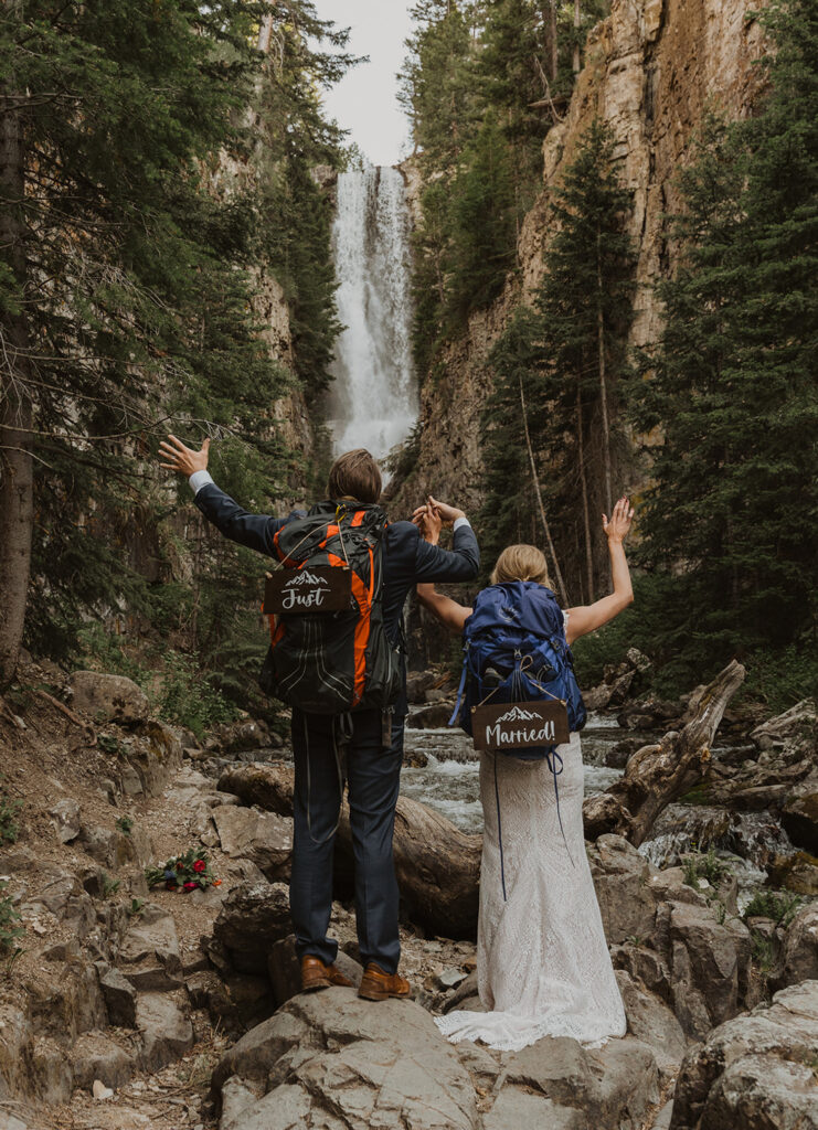 A couple wearing wedding attire and hiking gear, with "Just Married" signs after finding a national park to elope in!