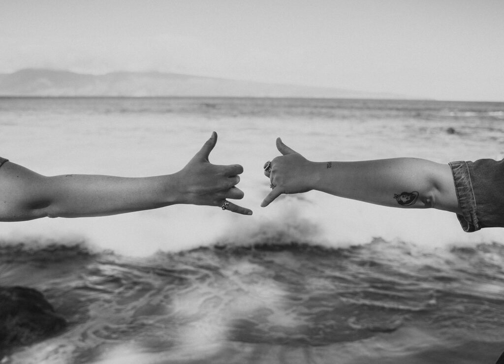 A same sex wedding couple elope in hawaii and throw up the hang loose sign on the beach. 